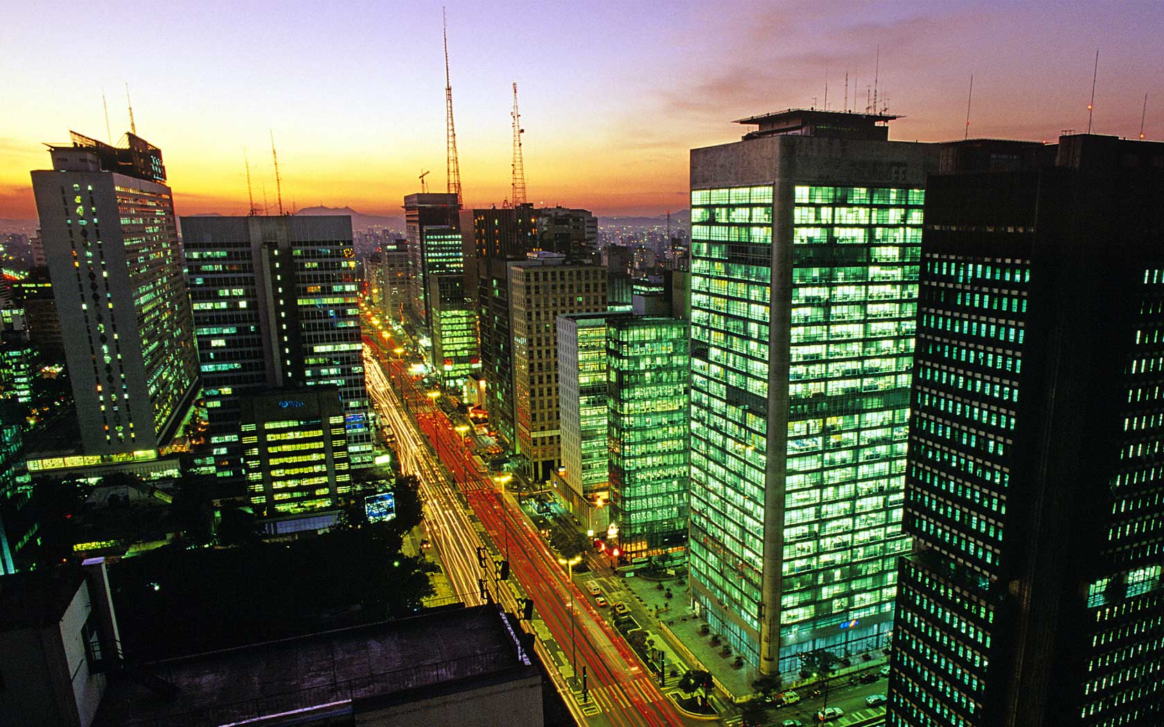 Avenida Paulista vista do alto ao anoitecer, com o céu alaranjado e os prédios iluminados